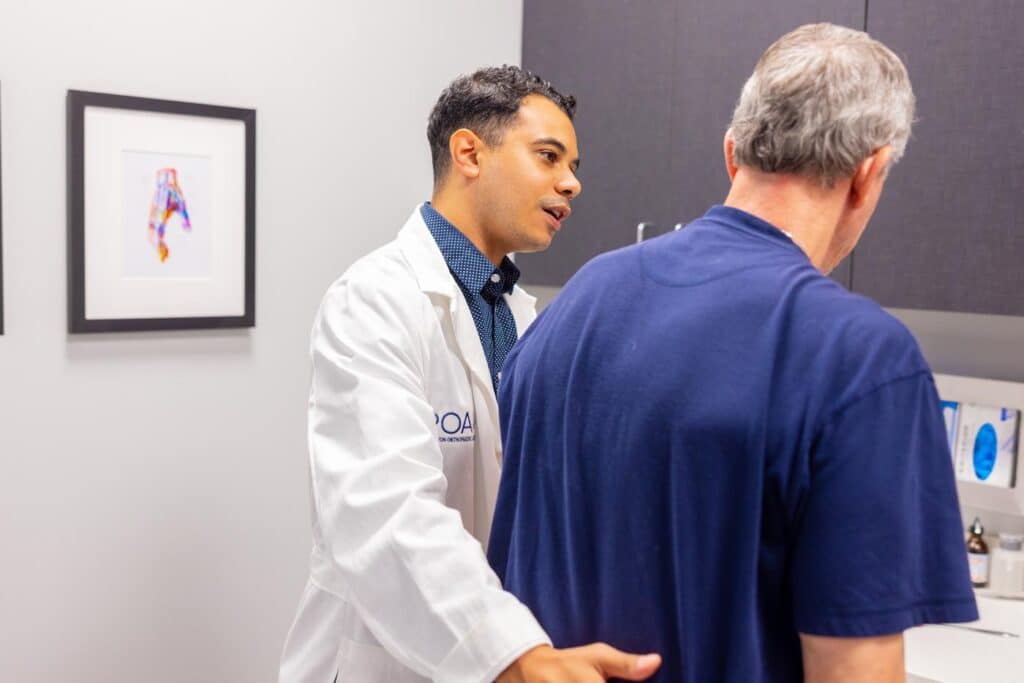 Dr. Saleh from Princeton Orthopaedic Associates stands right of center facing a patient, who is back to the camera, with his hand on the patients back, examining his spine.