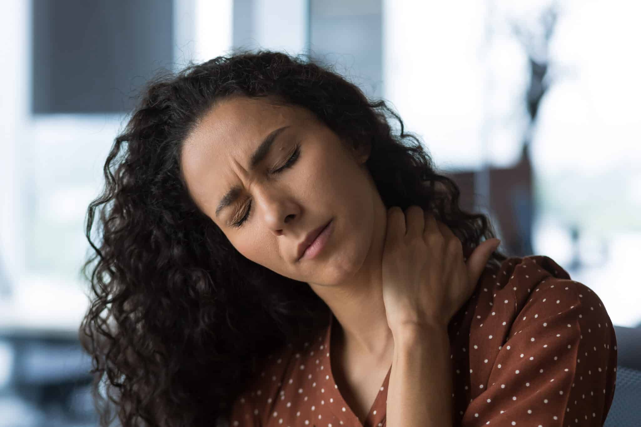 Close-up photo of curly-haired woman at home near the window with severe neck pain - Princeton Orthopaedic Associates woman sitting at desk tilting head holding her neck, in a neck stretch, with her left hand with an expression of pain.