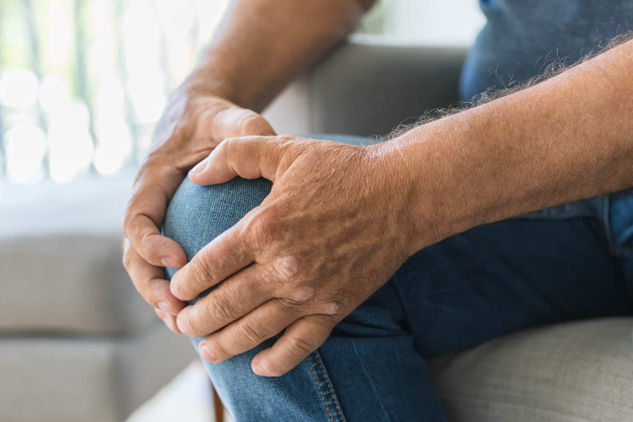 Close-up of weathered hands resting on a knee in blue jeans, indoors, with a soft blurred background
