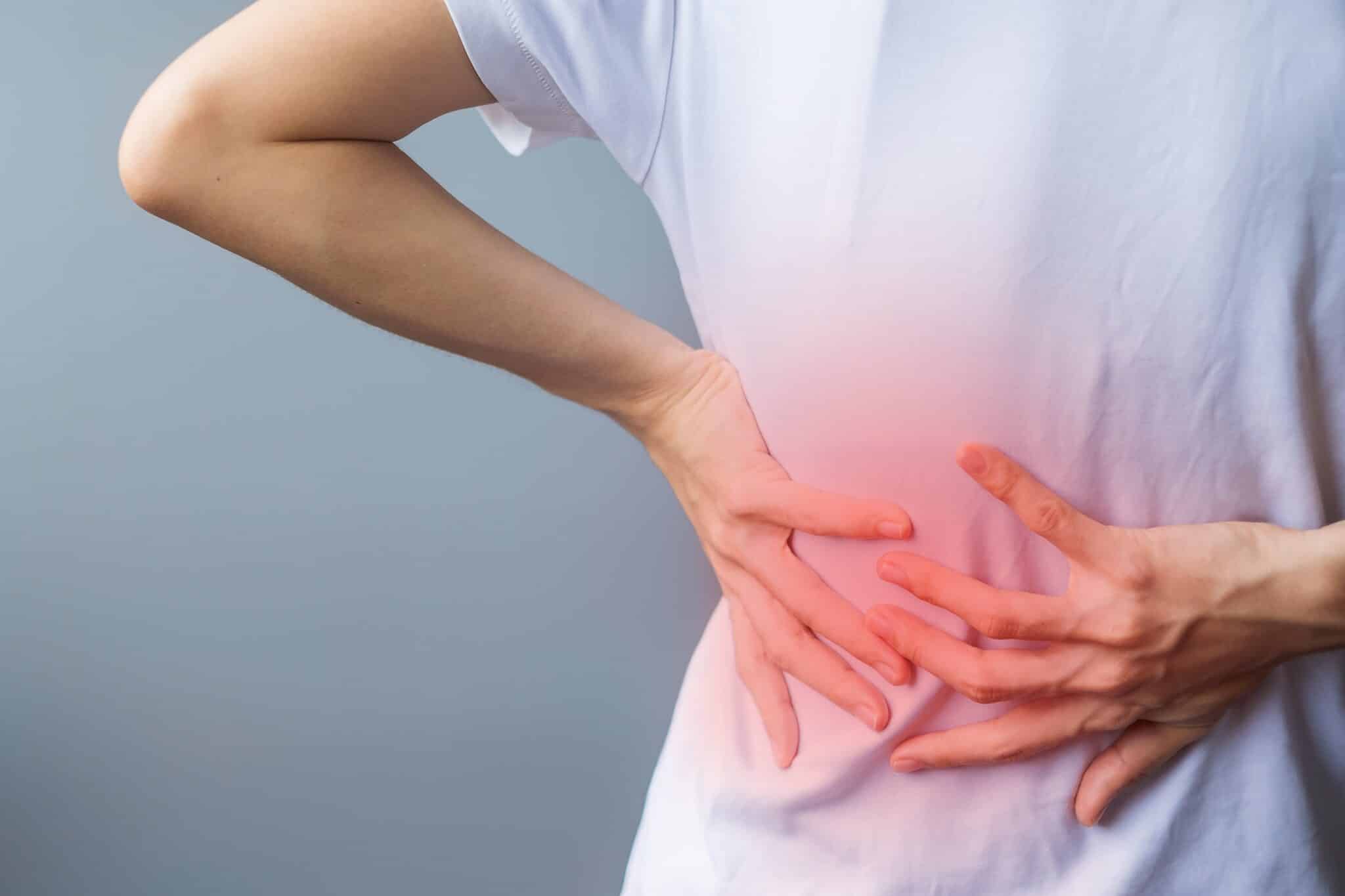 adult female with muscle pain on gray background Elderly woman - Princeton Orthopaedic Associates Person in a white shirt holding their midsection with both hands, signaling abdominal discomfort or cramps.