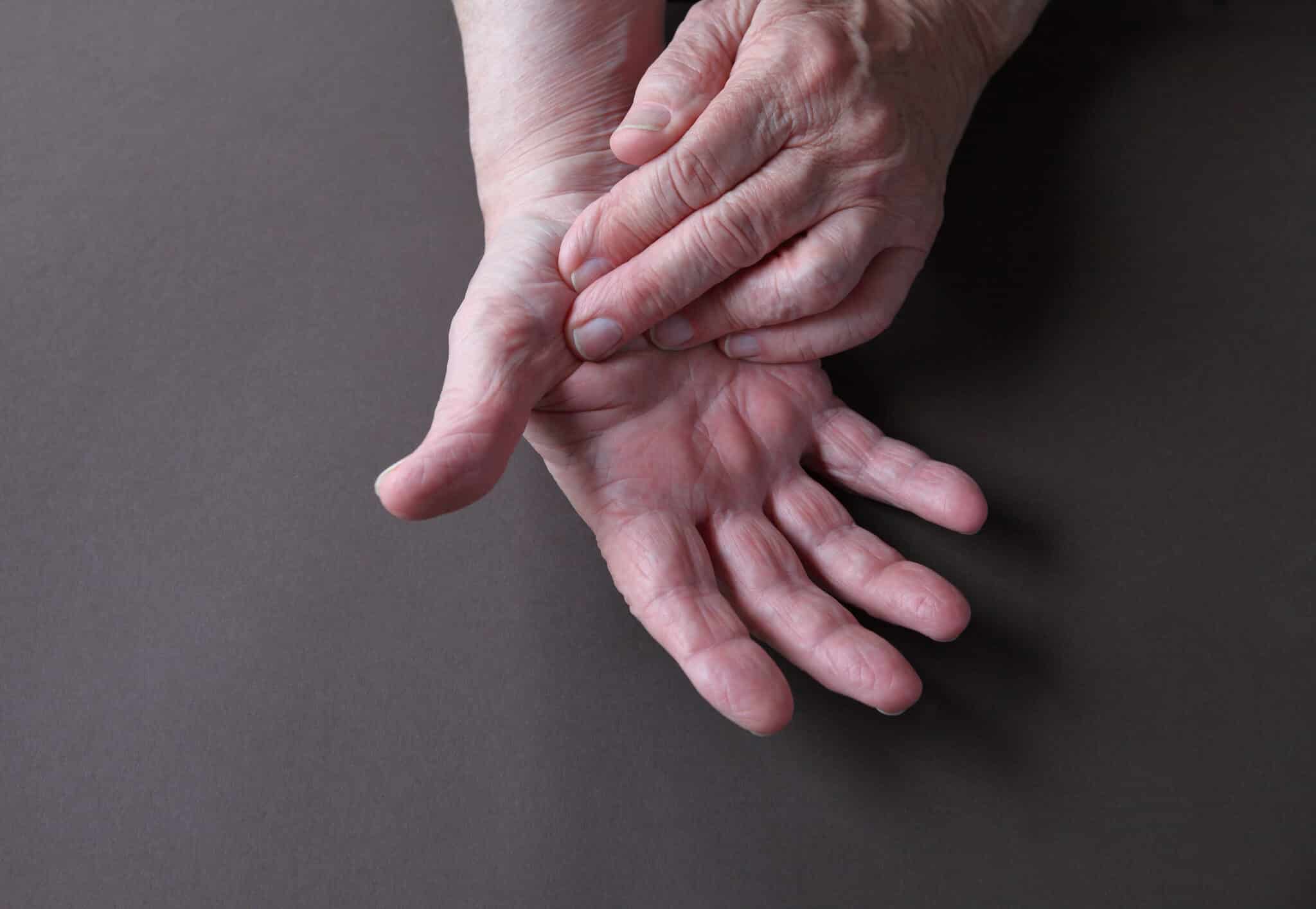 an-older-man-indicates-an-area-of-pain-on-his-thum-2026-03-26-06-22-00-utc - Princeton Orthopaedic Associates Close-up of elderly hands gently clasped, interlaced fingers against a gray background.