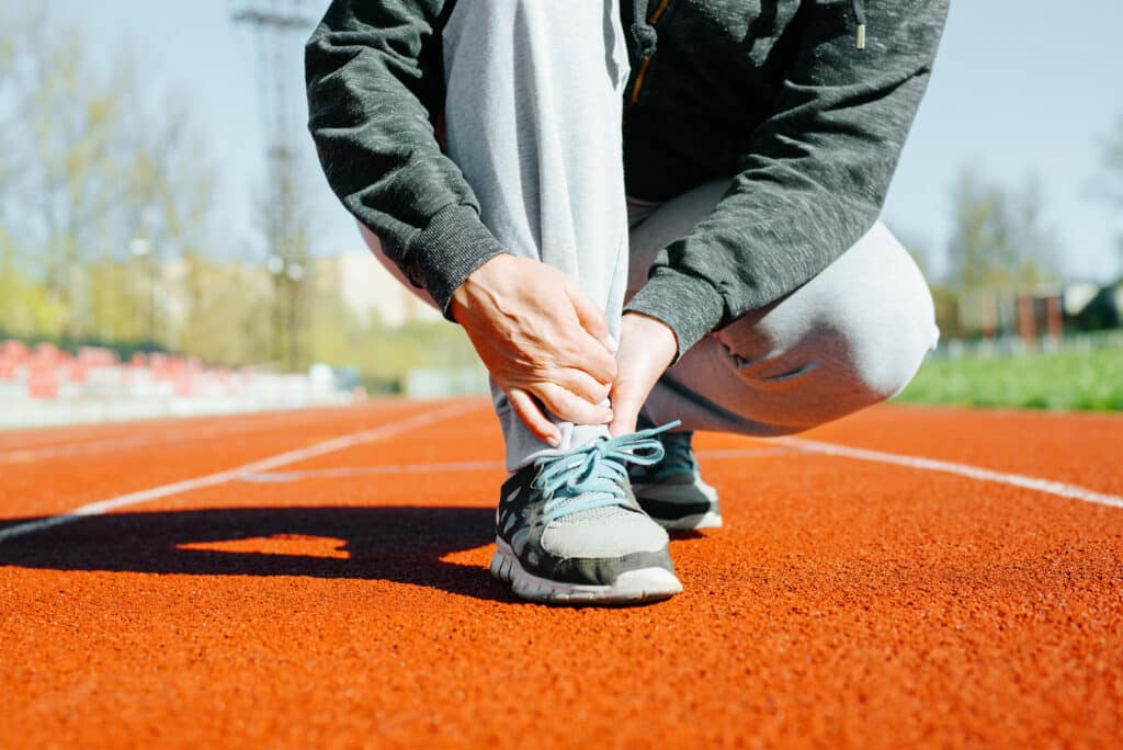 Sports injury while exercising outdoors Close-up of woman suffering from pain in tendons of lower leg and foot muscle sprain due to intense running training in stadium - Princeton Orthopaedic Associates Person tying light blue athletic shoes on a red track surface, close-up of hands and laces.