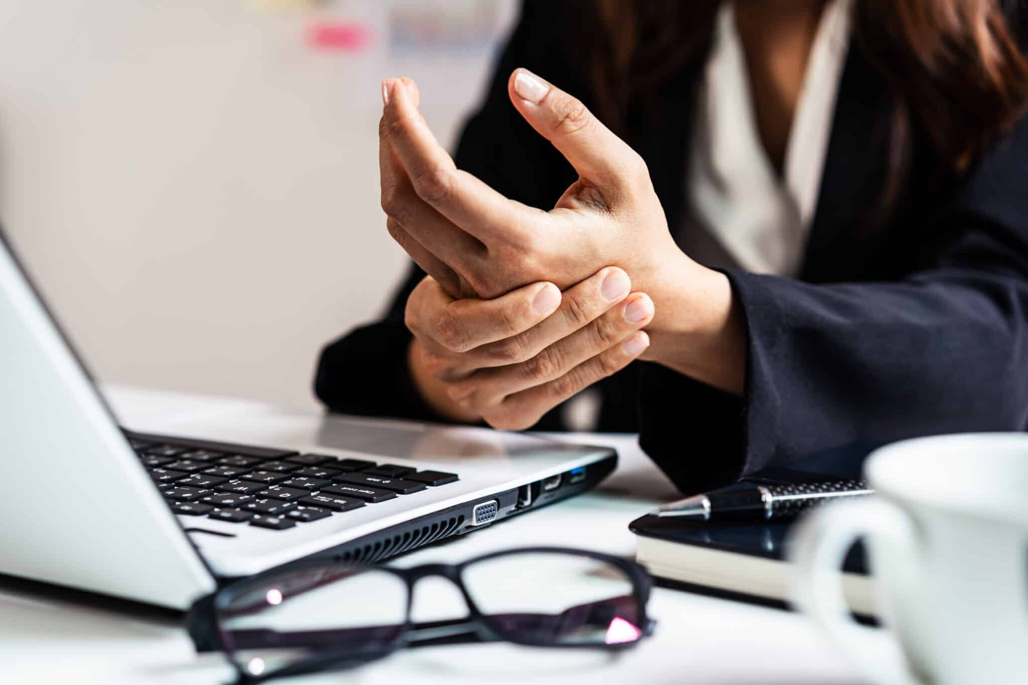 Stressed business woman suffering from wrist pain Office syndrome concept - Princeton Orthopaedic Associates Hands clasped over a desk near an open laptop, with glasses and a notebook in view.