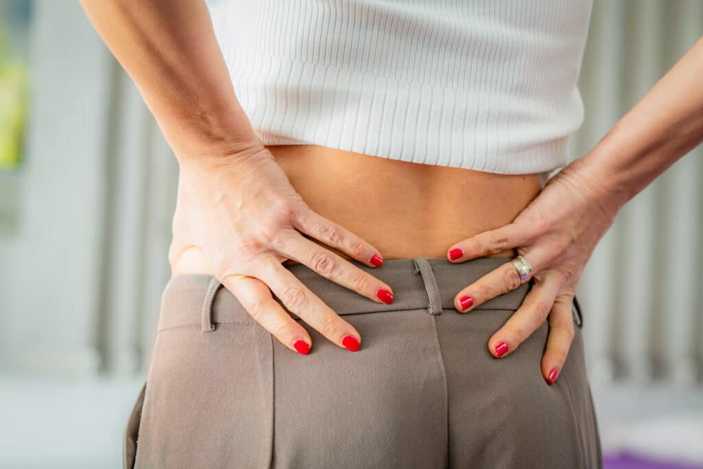 Woman experiencing lower back pain holding or massaging her sore back with discomfort The image highlights muscle strain lumbar pain sciatica arthritis or posture-related back issues - Princeton Orthopaedic Associates Hands resting on hips at waist, wearing a white ribbed top and gray pants, red nail polish visible. (informative)