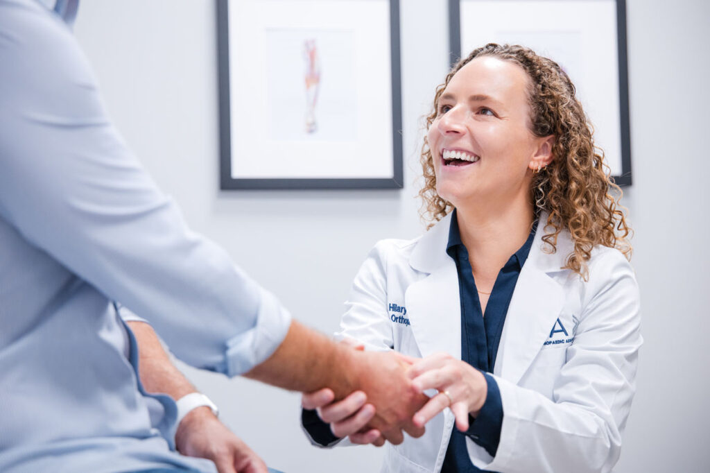Princeton Orthopaedic Associates - 2026 - Dr Campell-8 - Princeton Orthopaedic Associates Female doctor in a white coat smiles while shaking hands with a patient in a clinic.
