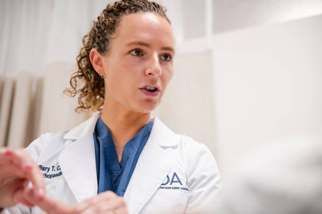 Princeton Orthopaedic Associates - 2026 - New Jersey - Dr Campbell-11 - Princeton Orthopaedic Associates Female healthcare professional in a white coat and blue scrubs speaking, in a clinical setting.