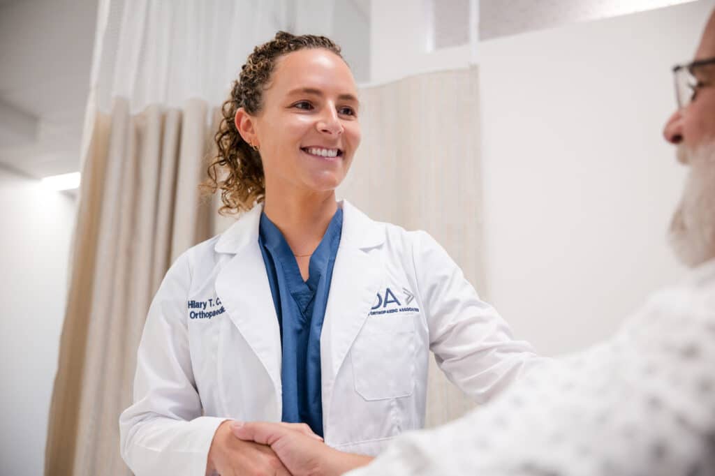 Princeton Orthopaedic Associates - 2026 - New Jersey - Dr Campbell-12 - Princeton Orthopaedic Associates Orthopedic doctor smiles and shakes hands with a patient in a clinic, wearing a white lab coat and blue scrubs.