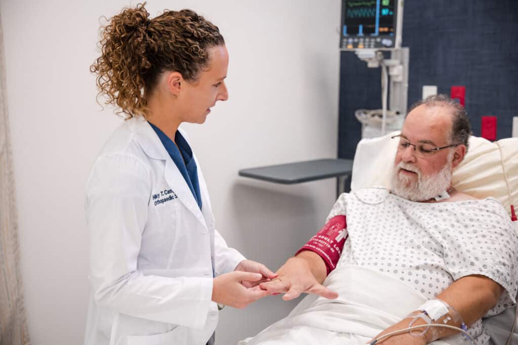 Princeton Orthopaedic Associates - 2026 - New Jersey - Dr Campbell-2 - Princeton Orthopaedic Associates Female doctor in a white coat gently holding an elderly patient's hand as he lies in a hospital bed with monitoring equipment nearby.