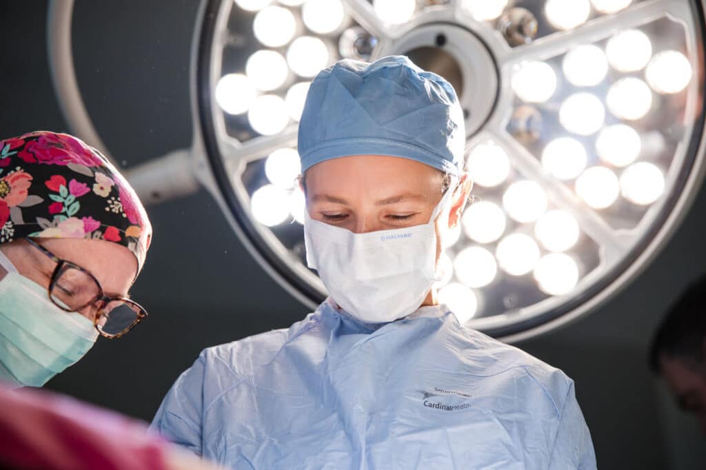 Surgeons in an operating room under bright circular lights; center surgeon in blue scrubs and mask preparing for surgery, Cardinal Health logo visible on gown.