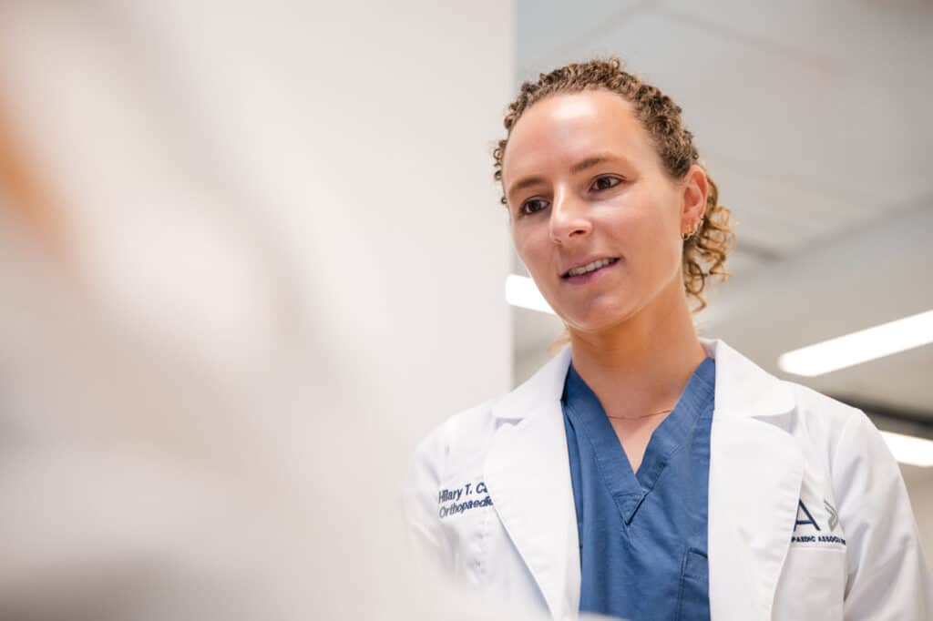 Female healthcare professional in a white lab coat over blue scrubs, smiling as she looks to the side in a clinical setting.