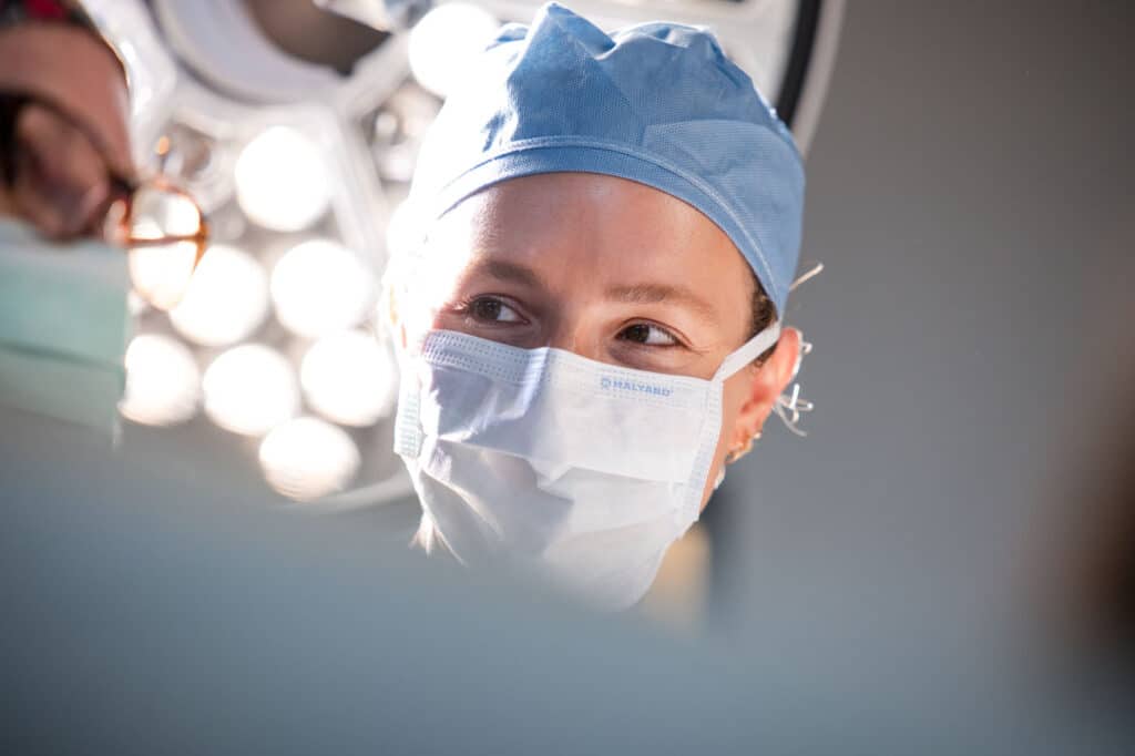 Princeton Orthopaedic Associates - 2026 - Surgical Center New Jersey -25 - Princeton Orthopaedic Associates Surgeon wearing a surgical mask and blue cap in an operating room, looking to the side with bright lights behind.