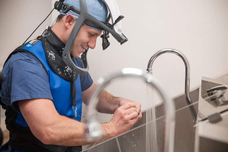 Male dental professional wearing blue scrubs and a protective headpiece washing his hands at a dental sink