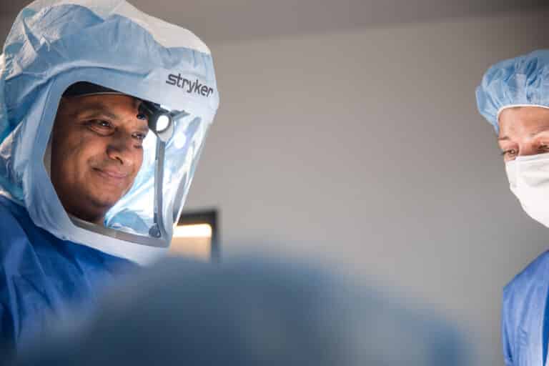 Two surgeons in blue scrubs and caps, wearing masks, performing an operation with headlamp illumination.