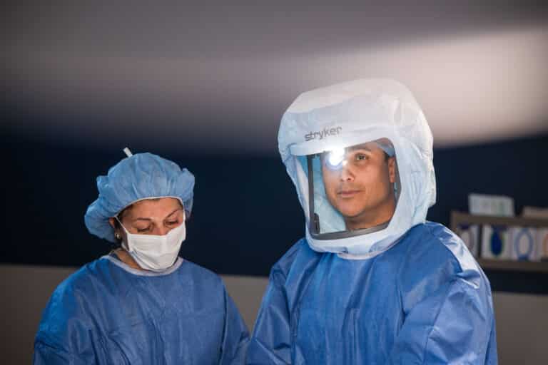 Two medical professionals in blue surgical gowns and caps, masks on, one wearing a headlamp and looking toward the camera in a clinic room.