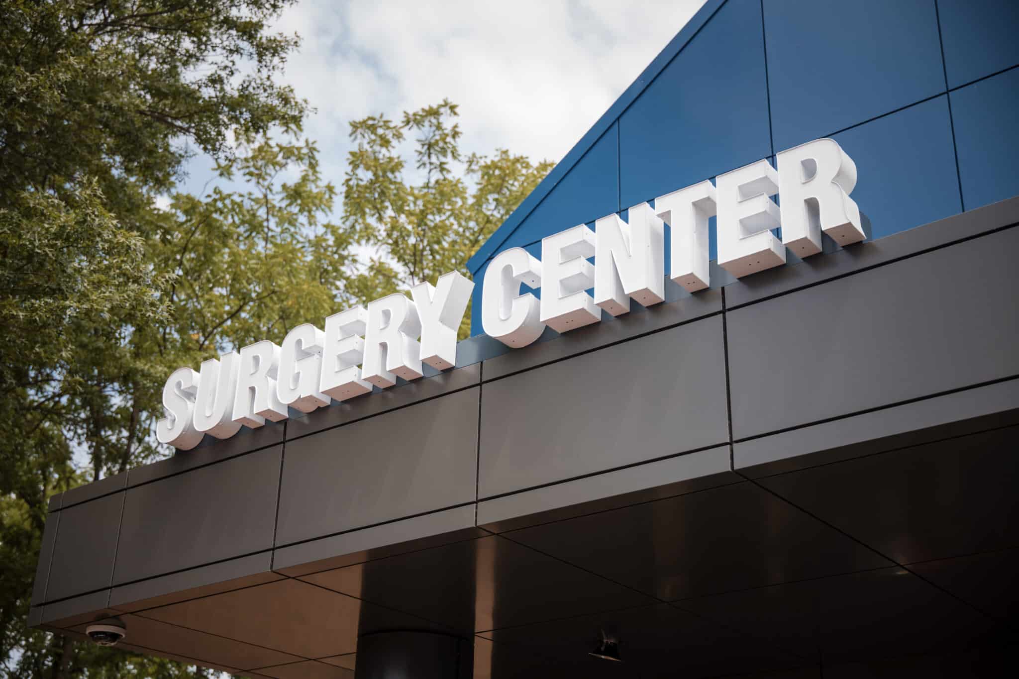 Side of a modern building with large white 3D letters spelling 'SURGERY CENTER' on a blue panel, trees in the background.