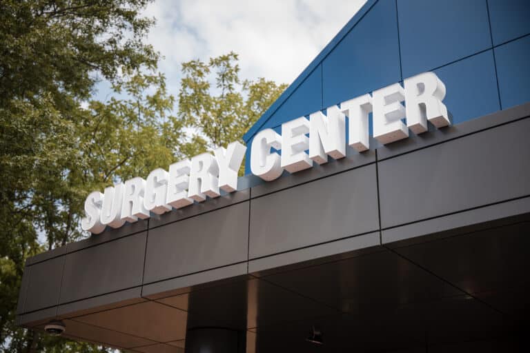 Exterior view of a building with a large white 'SURGERY CENTER' sign on a blue-panel facade, with trees in the background.