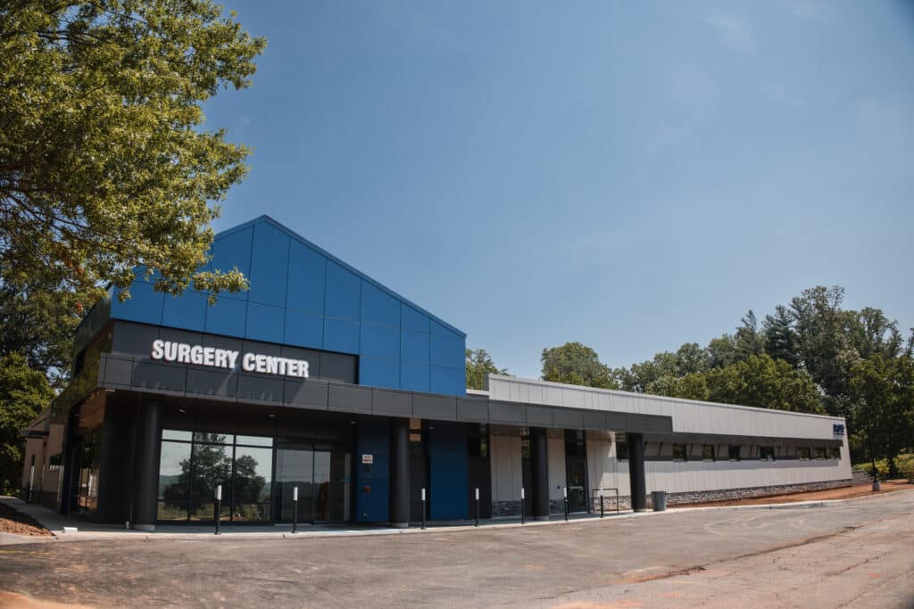 Princeton Orthopaedic Associates - 2026 - New Jersey - Front Desk-14 - Princeton Orthopaedic Associates Modern medical building with blue glass panels and a prominent 'SURGERY CENTER' sign, set against a sunny sky with trees nearby.