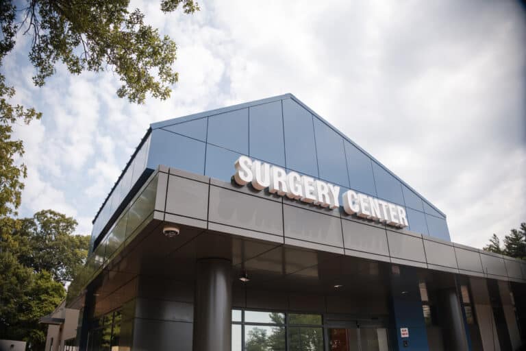 Exterior view of a modern glass building with the words 'SURGERY CENTER' on the facade, under a cloudy blue sky with trees surrounding it.