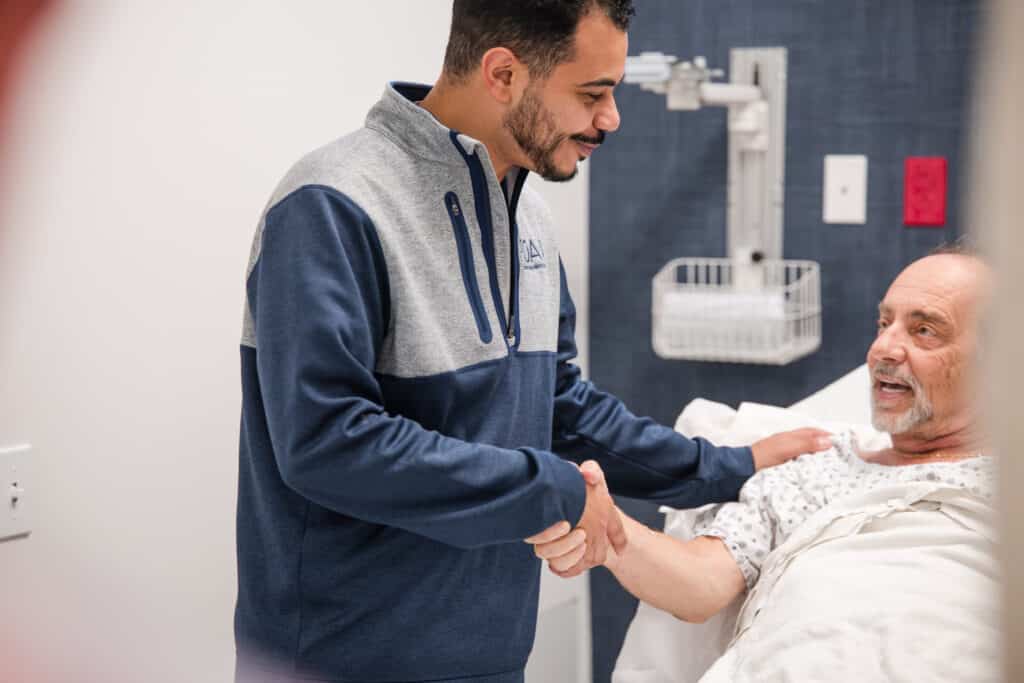 Caregiver in a blue-gray jacket holds an elderly man's hand at the bedside in a hospital room.