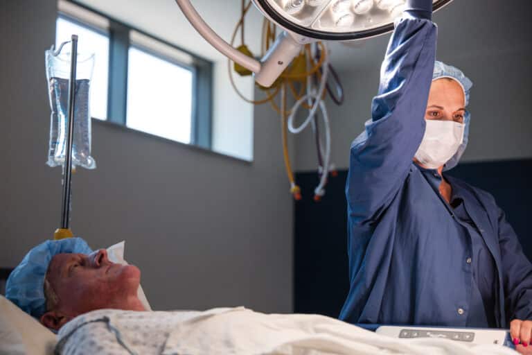 Surgeon or nurse in blue scrubs adjusts an overhead surgical light above a patient lying in bed.