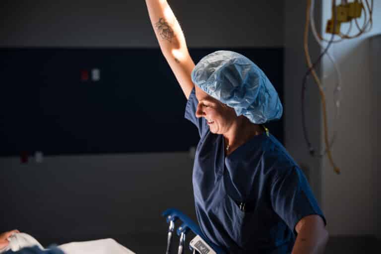 Nurse in blue scrubs and a hair cover raises an arm while smiling beside a patient in a hospital room.