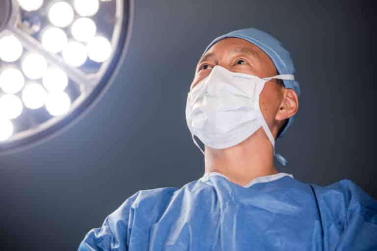 Surgeon in blue scrubs and a mask looks upward at bright overhead surgical lights in an operating room.