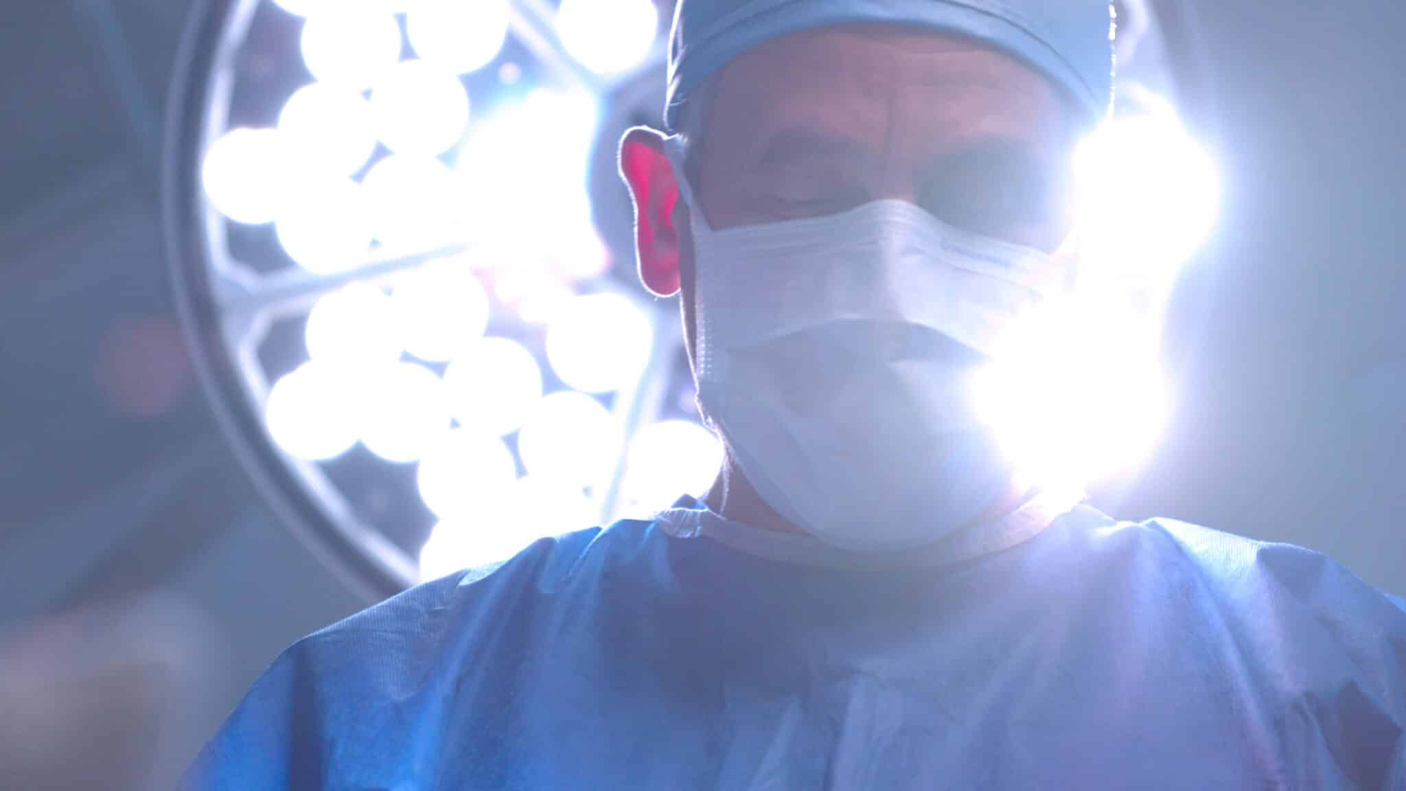 Surgeon wearing scrubs, mask and cap, viewed from behind in a brightly lit operating room